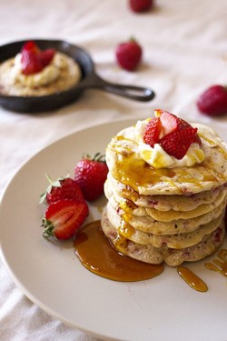 A stack of pancakes with strawberries and whipped cream on top with another small black pan of a pancake in the background
